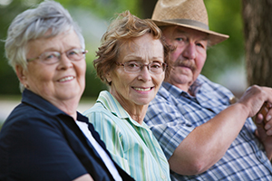 Two senior women both wearing glasses and a senior man wearing a hat sitting outside smiling